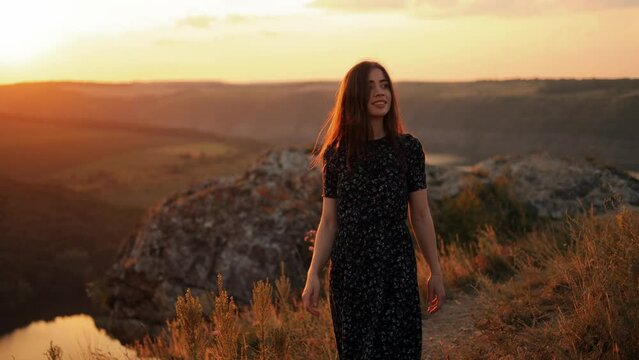 Portrait of young smiling beautiful woman in long dress walking on grass on hill with incredible view at sunset, outdoor walk, summer vacation