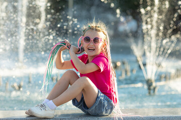 A little girl in a bright pink summer t-shirt with colored pink-blue African braids in her hair sits by the fountain and looks at camera. The child has a rest near a fountain during a heat wave.