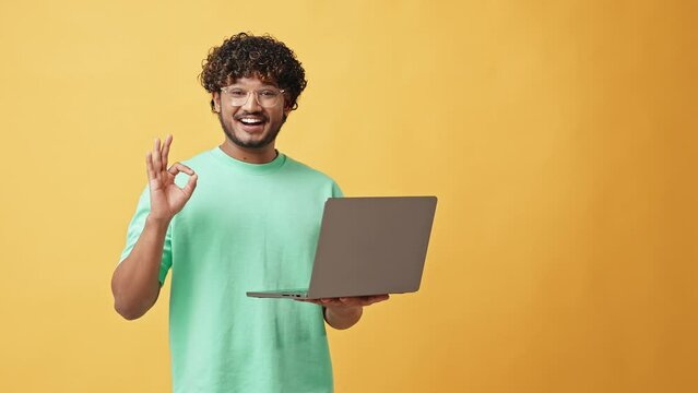 Adult stylish curly modern Indian man in glasses and turquoise t-shirt standing in studio on yellow background holding laptop and pointing finger at it and thumbs up gesture. - Powered by Adobe