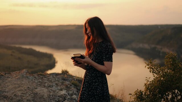 Young woman reading a book at sunset. A girl reads the Bible in the open air while standing on a rock, studying the word of God at sunset on top of a mountain. Searching for truth in the Holy Scriptur