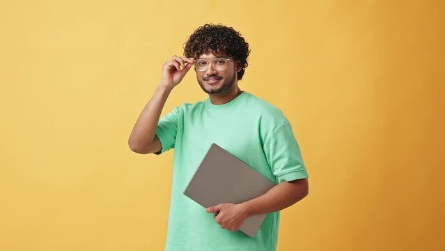 Adult stylish modern Indian man in glasses and turquoise t-shirt standing in studio on yellow background holding laptop and adjusting glasses looking at camera.
