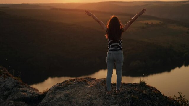 Silhouette of a woman hiker standing alone on big stone at sunset in mountains. Female tourist raising her hands up on high rock in evening nature. Tourism, traveling and healthy lifestyle concept.