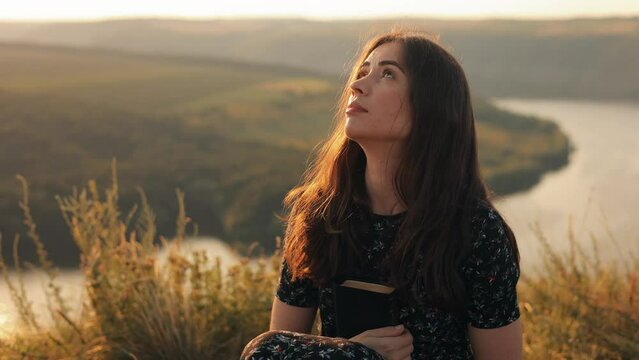  Beautiful young woman reading the bible sitting on top of a hill with a beautiful view at sunset, admiring the view and nature, relaxing alone with thoughts, slow motion
