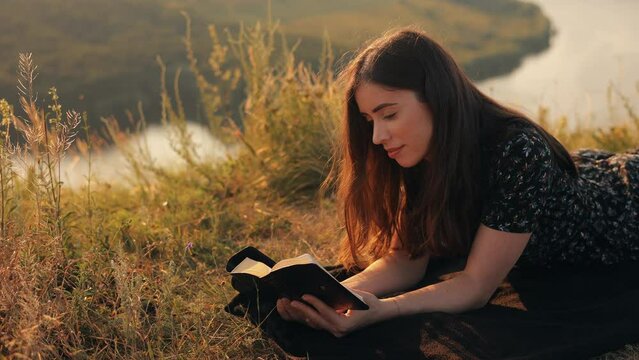 Young christian girl reading the bible, praying, putting her hand on the bible, lying in the grass on the rock in nature with an incredible view, sunset, peace and tranquility, faith