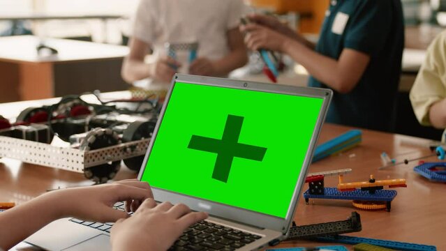 Child sitting at the table and writing the code for the moving robot on a laptop with a mockup green screen close up in the back view in the class at the robotics lesson. Concept of technological