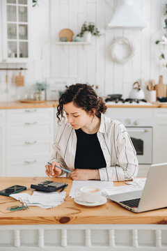 Focused caucasian woman working at home in the kitchen with financial papers, counting on a calculator, paying bills, planning a budget to find a way to save money. Self-employed accounting