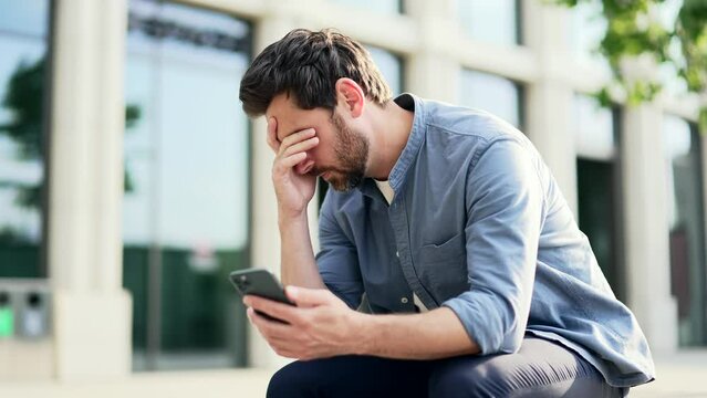 Depressed employee entrepreneur, freelancer sitting on bench near office center upset looking at mobile phone smartphone reading bad news or negative result. Sorrowful worker suffering from depression