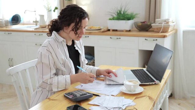 Focused caucasian woman working at home in the kitchen with financial papers, counting on a calculator, paying bills, planning a budget to find a way to save money. Self-employed accounting