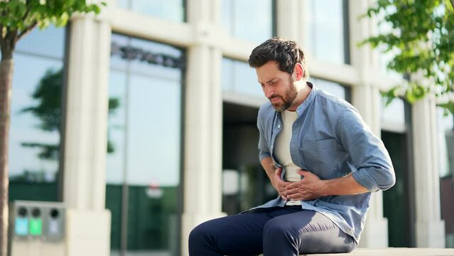 Business man suffering severe stomach pain nausea or poisoning sitting on bench on city street outside outdoors. Mature employee adult male office worker suffering from indigestion abdominal ache