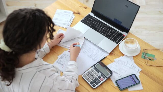 Top view of a woman working at home in the kitchen with financial papers, counting on a calculator, paying bills, planning a budget to save some money. Independent accounting, remote accountant