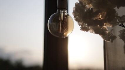 Close up hanging electric light bulb and arrangement of pastel colours roses flowers by the window with sunset sunlights. Stylish details of festive decorations indoors restaurant - Powered by Adobe