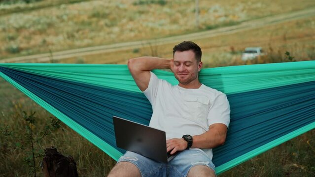 Joyful Caucasian man sits in hammock with laptop on his knees. Relaxed man working distantly away from office.