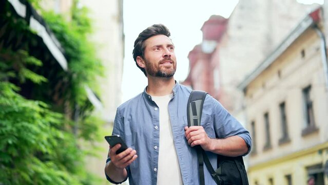 Handsome bearded man, a tourist with backpack, walk explore city street, using mobile phone. Happy male tourist traveling browsing smartphone looking at navigation app or map. Traveller lifestyle