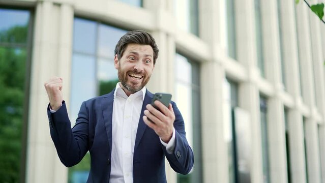 Happy businessman investor is happy reading receive great good news on mobile phone. Business man in formal suit celebrates victory look at smartphone Standing near office building on a city street