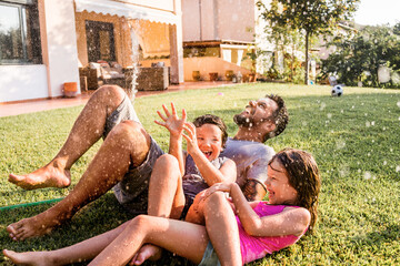 Father playing with his kids in the backyard with a water hose - obrazy, fototapety, plakaty