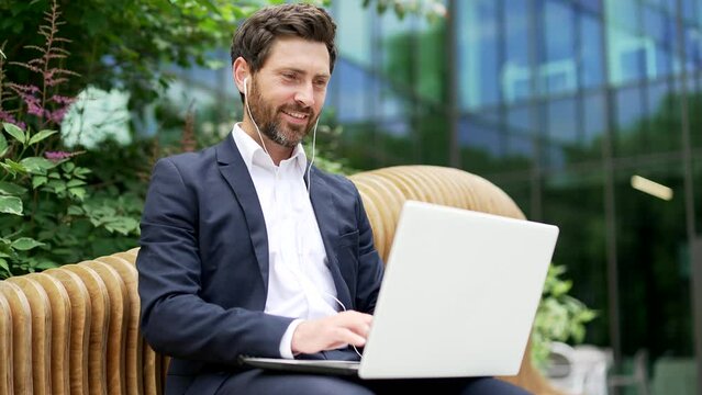 Handsome bearded businessman in suit working with laptop sitting on bench near modern office business center Happy Employee Executive typing browse laptop outside outdoors in city street Closeup smile