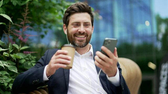 Relaxed businessman rest using mobile cell phone browsing watching social media sitting outside during break with coffee cup in hands. Outdoors Business man on a bench near office building. Lifestyle