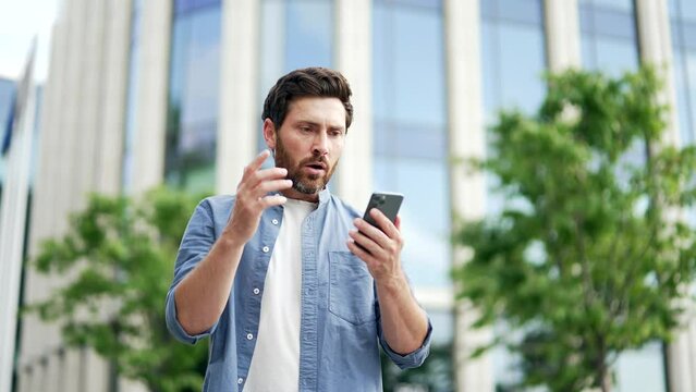 Mature adult businessman receives bad news reading sms message looking at smartphone phone screen standing near modern business center on city street outdoors outside. Frustrated sad man