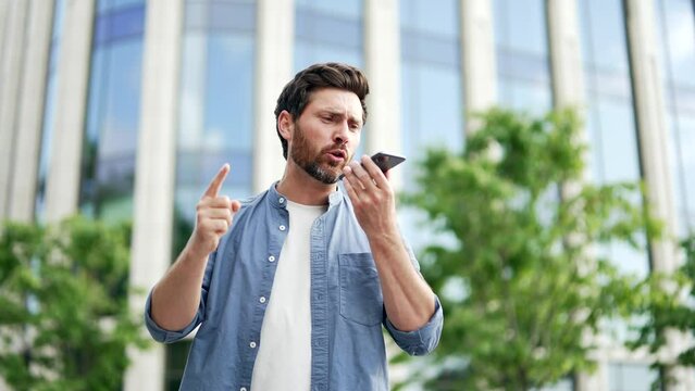 Angry businessman talking emotionally while holding smartphone while standing outside. Shirty irate mature male manager entrepreneur in casual arguing on the phone in front of an office building