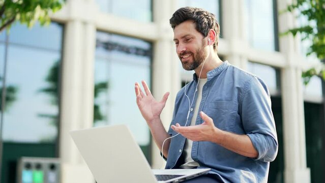 Businessman communicates remotely on a video call while sitting on a bench near a modern office center outside the building. Bearded business male entrepreneur in casual talking online meeting Outdoor
