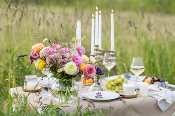 Romantic table decor for a loving couple on a meadow. Two glasses of wine, flowers in a vase, silverware, fruits, chandelier with burning candles, wooden furniture. Sunset, golden hour, summer