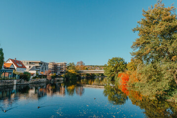 Beautiful small river with clean and clear water front of colorful autumn trees and small old town on the hill agaist nice blue and clouds sky during autumn in Europe