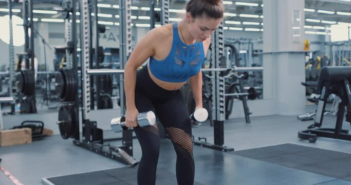 Fit serious motivated woman in sportswear bending forward and lifting dumbbells, equipment on background. female athlete working out in gym. back workout Slow motion Side view