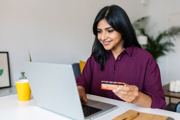 Smiling young indian woman holding credit card and using laptop computer to do online shopping