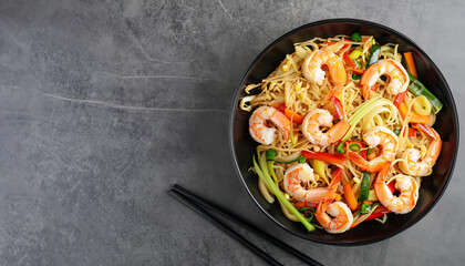 Stir fry noodles with vegetables and shrimps in black bowl. Slate background. Top view. Copy space.