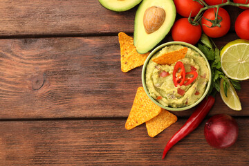 Bowl of delicious guacamole, nachos chips and ingredients on wooden table, flat lay. Space for text