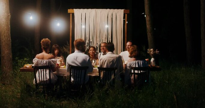 The bride and groom embrace sitting at the festive table next to their guests. A man takes pictures of the bride and groom on his mobile. Outdoor wedding feast