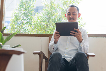 Young business man using tablet sitting at the chair in a home , communicating online, distantly working or studying on tablet at home.