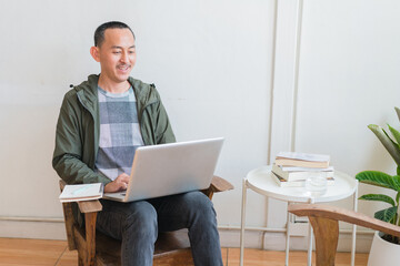 Young business man using laptop sitting at the chair in a home , looking at the paper, communicating online, writing emails, distantly working or studying on computer at home.