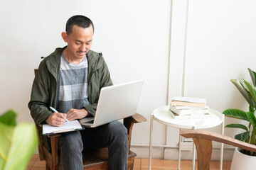 Young business man using laptop sitting at the chair in a home , looking at the paper, communicating online, writing emails, distantly working or studying on computer at home.