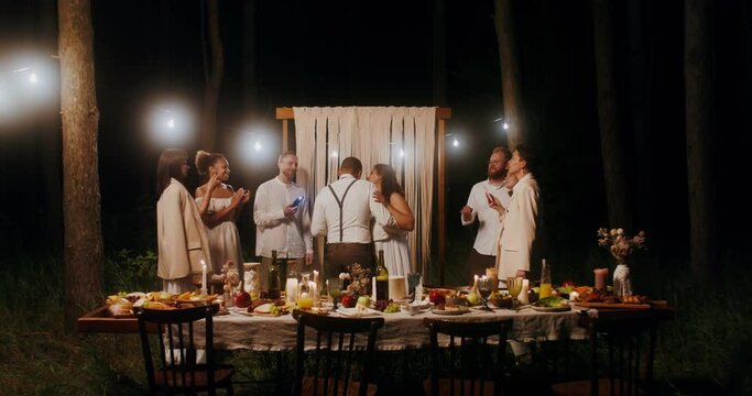 The bride and groom are dancing surrounded by their friends at an outdoor wedding feast at night