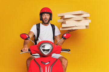 Young male courier wearing red helmet in a hurry riding red scooter and holding pizza boxes in one hand, fast delivery concept, copy space