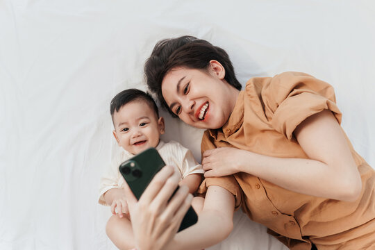 Happy Asian Mother And Her Newborn Baby Making A Selfie Or Video Call To Father Or Relatives In A Bed. Concept Of Technology, New Generation,family, Connection, Parenthood