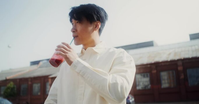 A young man with a paper bag in his hands drinks juice from a disposable cup with a straw while walking around the city