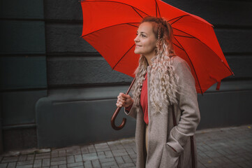 Mature blonde woman under a red umbrella on the street. trendy wearing middle aged woman with fashion hair walking in the city