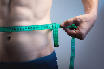 Torso of a young athletic man holding a measuring tape around his waist, on a gray background. a...