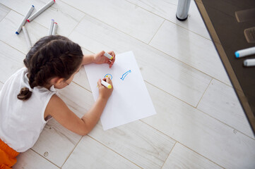 View from above preschooler girl drawing picture on white paper sheet with watercolor...
