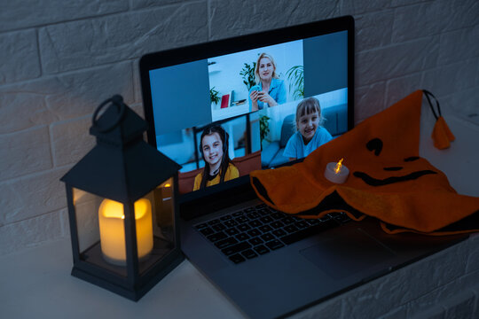 Group Of People Happy To See Each Other At Virtual Halloween Party. Laptop Computer At Decorated Kitchen Table, Waving Hand And Greeting Friends At Online Video Meeting