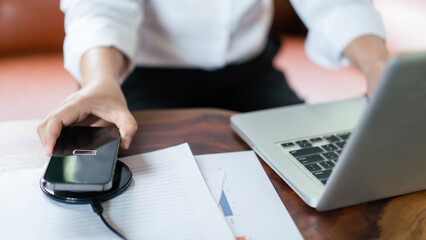 Hands of businessman charging battery smartphone on wireless charger device and type data on laptop