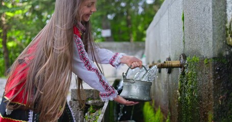 Bulgarian girl in traditional folklore attire, ethnic slavic clothing pouring water from old stone tap or fountain in Bulgaria. Culture, tradition and lifestyle in Bulgaria