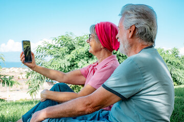 Selfies concept. Happy senior couple sitting in meadow using smartphone to take selfie, smiling grey-haired man and woman looking into phone camera