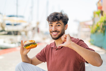 Young Arabian handsome man holding a burger at outdoors proud and self-satisfied