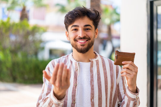 Handsome Arab man holding a wallet at outdoors inviting to come with hand. Happy that you came