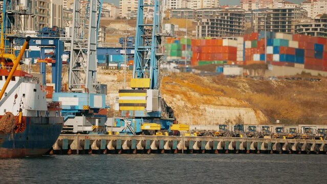 4K scene from commercial port in daylight. Forklift heading towards docked ship for unloading container with stacked containers and buildings in the background stock video. 