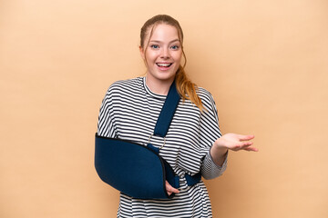 Young caucasian girl with broken arm and wearing a sling isolated on beige background with shocked...