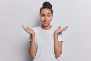 Waist up shot of confused Latin woman raises her hands with palms open conveying sense of hesitation doubt unable to make decision makes difficult choice wears casual t shirt isolated on white wall